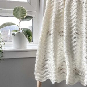 A small throw made with a gentle chevron crochet stitch shown hung over a blanket ladder, by a window with a plant on the sill